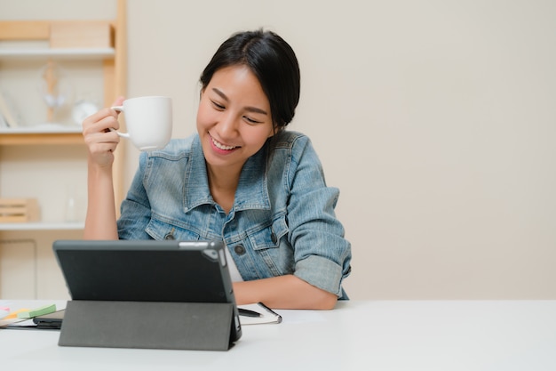 Mulher asiática nova que trabalha usando a tabuleta que verifica meios sociais e que bebe o café quando relaxe na mesa na sala de visitas em casa.