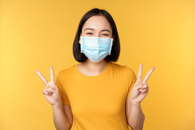 Mulher asiática positiva sorrindo usando máscara facial médica de covid19 durante pandemia mostrando gesto de paz vsign fundo amarelo