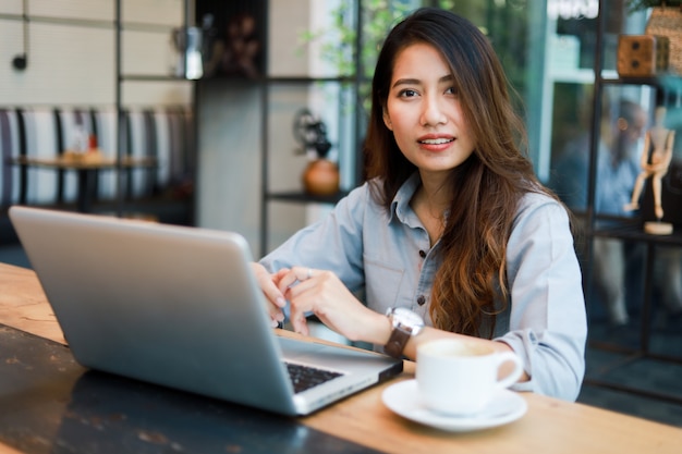 Mulher asiática que trabalha e bebe café no café com sorriso de computador portátil e trabalho feliz