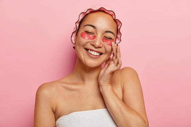 Mulher asiática satisfeita aplica adesivos de colágeno para reduzir o inchaço, sorri positivamente, mostra dentes brancos, usa touca de banho macia, fica enrolada em toalha, faz procedimentos de beleza antes do encontro com o namorado