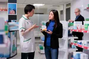 Foto grátis mulher asiática segurando pacote de vitaminas e ouvindo farmacêutico explicando instruções de dosagem de ácido ascórbico. cliente de farmácia comprando suplemento alimentar, conversando com assistente de farmácia no corredor