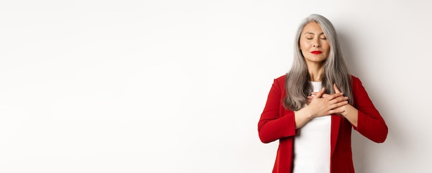 Foto grátis mulher asiática sênior com lábios vermelhos e blazer fecha os olhos e segurando as mãos no coração sentimento de gratidão n