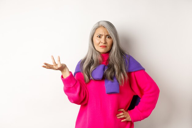 Mulher asiática sênior zangada e confusa com as mãos estendidas para o lado e olhando para a câmera, perplexa, usando um suéter rosa contra um fundo branco