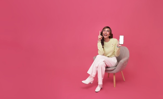 Mulher asiática sentada no sofá sentindo felicidade e gesto mostrando a tela em branco do smartphone móvel