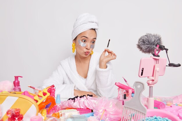Mulher asiática séria blogueira de beleza grava vídeo para seu vlog de beleza segura escova cosmética aplica tapa-olhos fala sobre procedimentos de cuidados com a pele faz maquiagem em casa na frente da câmera do smartphone