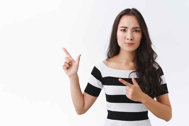 Mulher asiática séria cética em camiseta listrada parece suspeita e insegura sobre a oferta duvidosa apontando o canto superior esquerdo franzindo a testa e apertando os olhos câmera expressa dúvidas e desacordo