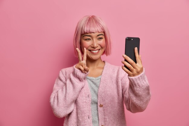 Mulher asiática sincera sorridente faz gesto de paz, dá as boas-vindas ao amigo durante videoconferência, tira selfie via smartphone, veste um macacão casual, tem o cabelo tingido da moda