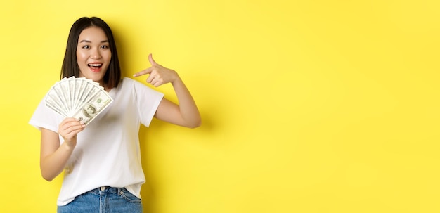 Mulher asiática sorridente apontando as mãos para dinheiro mostrando dólares em pé sobre fundo amarelo