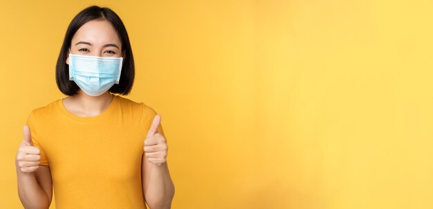 Mulher asiática sorridente com máscara facial médica mostrando os polegares para aprovação como e recomendando smth em pé