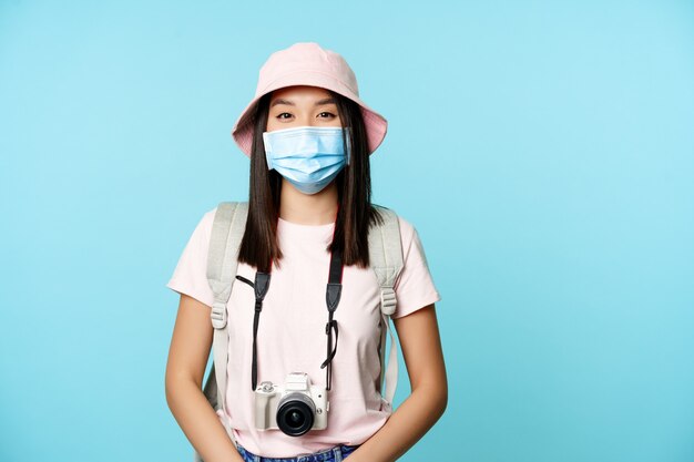 Mulher asiática sorridente com máscara médica viajando durante a pandemia em pé com a câmera fot ...