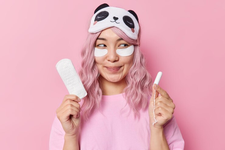 Foto grátis mulher asiática sorridente de cabelo rosa satisfeita tem expressão sonhadora aplica manchas de beleza sob os olhos para refrescar a pele escolhe o melhor produto de higiene segura absorvente higiênico e tampão tem menstruação