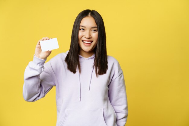 Mulher asiática sorridente de compras mostrando um cartão na mão em pé sobre um fundo amarelo