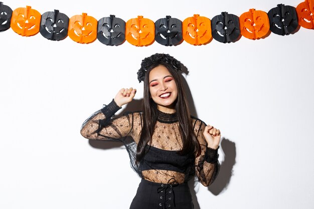 Mulher asiática sorridente despreocupada com fantasia de bruxa, aproveitando a festa de halloween, dançando e regozijando-se, em pé sobre um fundo branco com decoração de abóbora.