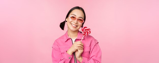 Mulher asiática sorridente em óculos de sol segurando doces de pirulito comendo doces e parecendo feliz em pé sobre fundo rosa