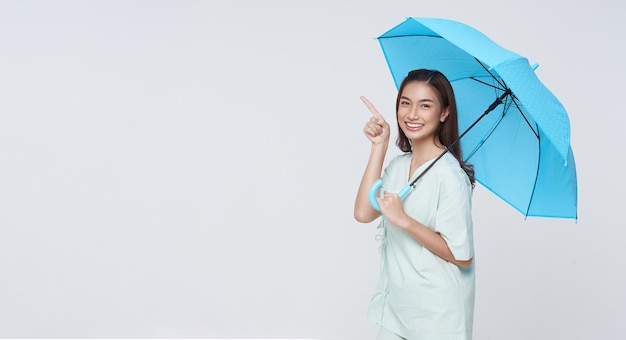 Mulher asiática sorridente feliz em pé e segurando o guarda-chuva azul e apontando o dedo para copiar o espaço