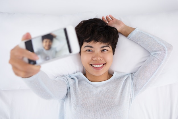 Mulher asiática sorridente que tira foto de Selfie na cama