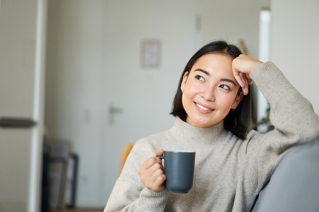 Mulher asiática sorridente sentada no sofá com sua caneca tomando café em casa e relaxando depois do trabalho parecendo calma e aconchegante