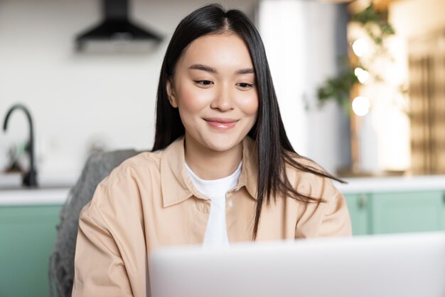 Mulher asiática sorridente usando laptop de menina em casa senta-se na cozinha com computador educação à distância ...