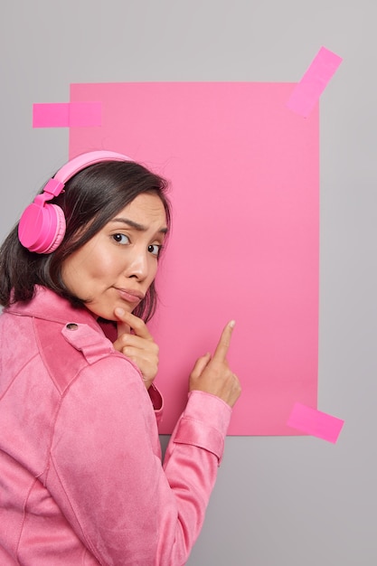 Mulher asiática triste e perplexa com cabelos escuros apontando para um espaço vazio para o seu conteúdo de publicidade sugere o uso de banner promocional para colocar suas informações ouve música em fones de ouvido sem fio