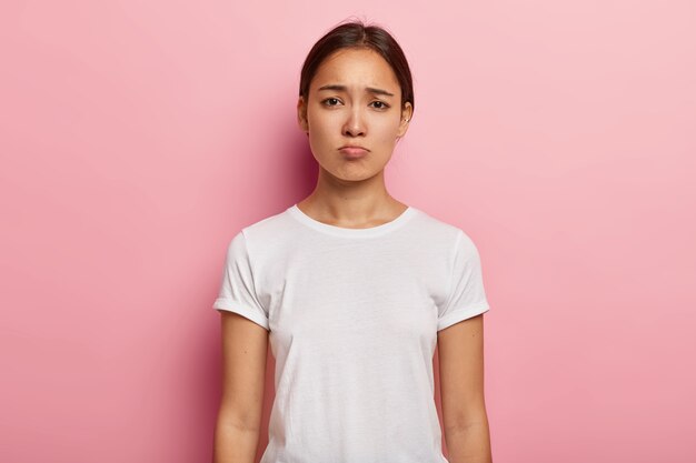 Mulher asiática triste e sombria tem olhar infeliz, expressa pesar e tristeza, franze a testa, fica frustrada, veste camiseta branca, mostra emoções negativas chateada por más notícias, reclama sobre vida injusta