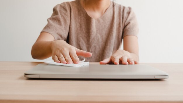 Mulher asiática usando spray de álcool no laptop limpo de tecido antes de abrir para proteger o coronavírus. Computador limpo feminino para higiene quando o distanciamento social fica em casa e no tempo de quarentena.