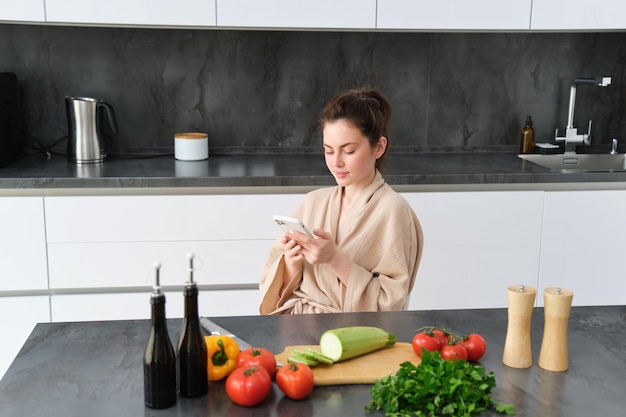 Mulher assistindo receita no smartphone sentada na cozinha com legumes e tábua de cortar