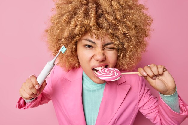 Mulher atenta com cabelo encaracolado morde doce de caramelo segura escova de dentes elétrica come alimentos prejudiciais aos dentes fala sobre efeito destrutivo ao comer açúcar e doces Maus hábitos para a saúde bucal