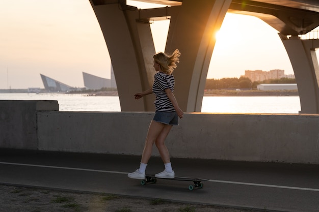 Mulher ativa aproveitando a noite de verão patinando em longboard ao longo do rio com vista para a cidade e o pôr do sol