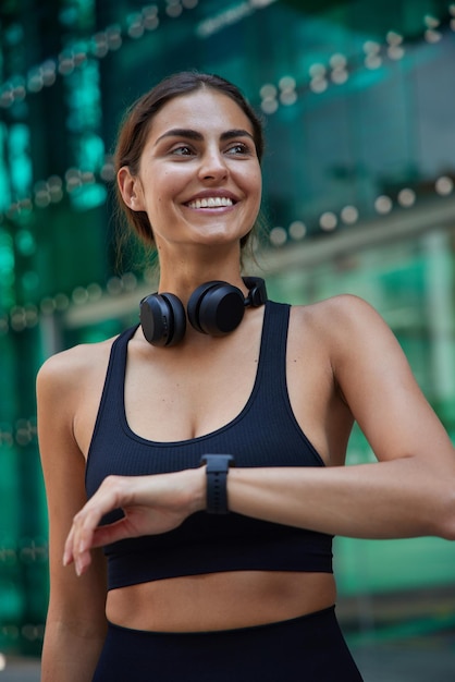 Mulher ativa esportiva com sorriso alegre no rosto verifica os resultados após o treino via smartwatch vestido com top cortado preto usa fones de ouvido no pescoço estando de bom humor modelo feminino atlético