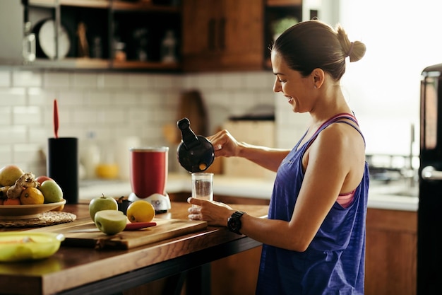 Mulher atlética feliz derramando smoothie de frutas em um copo na cozinha