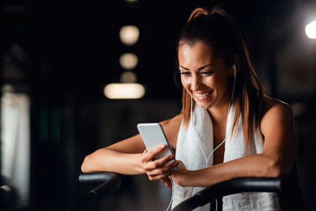 Mulher atlética feliz usando telefone inteligente e ouvindo música enquanto se exercita em uma academia