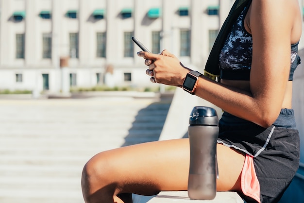 Mulher atlética usando seu telefone celular e relaxando depois do treino ao ar livre. Esporte e estilo de vida saudável.