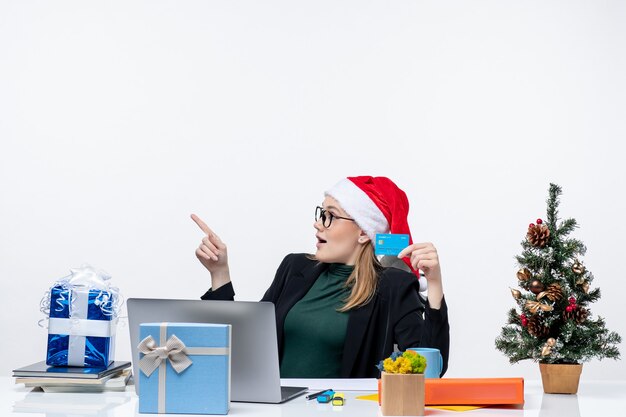 Mulher atraente com chapéu de papai noel e usando óculos, sentada à mesa de presente de natal e segurando o cartão do banco no escritório