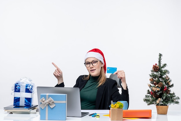 Mulher atraente com chapéu de papai noel e usando óculos, sentada em uma mesa de presente de natal e segurando um cartão do banco na imagem de estoque do escritório