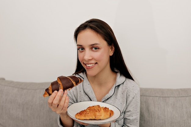 Mulher atraente, com longos cabelos escuros de pijama, tomando café da manhã em casa pela manhã. Ela segura um croissant, sorrindo