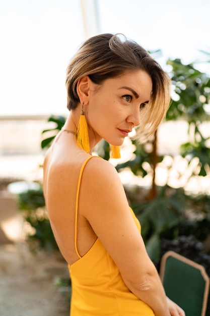 Mulher atraente com vestido amarelo de verão com penteado curto em estilo vintage boho posando no quarto interior