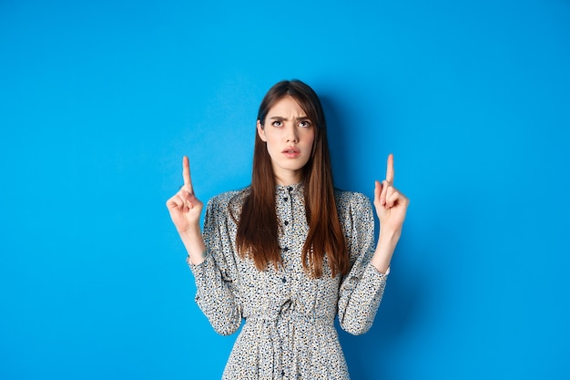 Mulher atraente confusa num vestido franzindo a testa apontando os dedos para cima e parecendo intrigada, não consigo entender ...