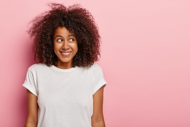 Mulher atraente de cabelos escuros encaracolados olha à parte, tem um sorriso encantador, está de bom humor após o encontro ou festa, usa camiseta casual maquete, fica de pé sobre uma parede rosada, espaço livre à parte. Pessoas e emoções