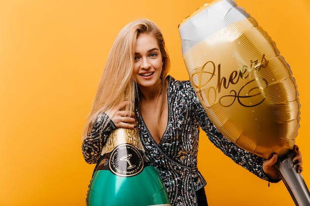 Mulher atraente de cabelos louros segurando a garrafa de champanhe. Mulher branca feliz se preparando para a festa de aniversário com um sorriso.