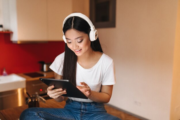 Mulher atraente de top branco curtindo música em fones de ouvido e segurando o tablet enquanto está sentado na cozinha