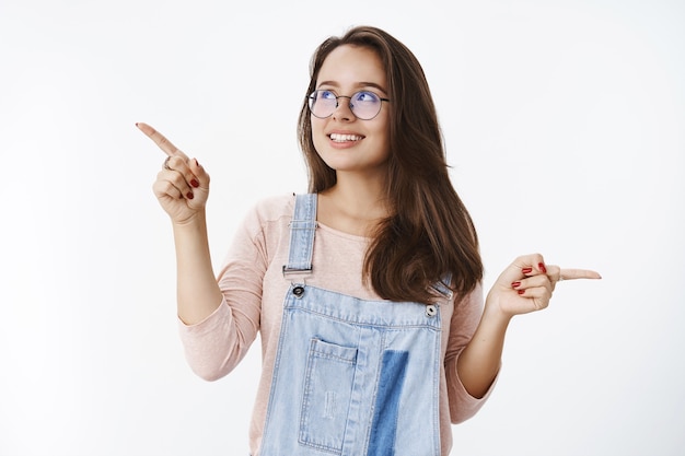 Mulher atraente dos anos 20 de óculos e macacão jeans escolhendo o produto com olhos sonhadores e sorriso largo olhando para o canto superior esquerdo apontando para o lado fazendo a escolha com muitas variantes sobre a parede cinza.