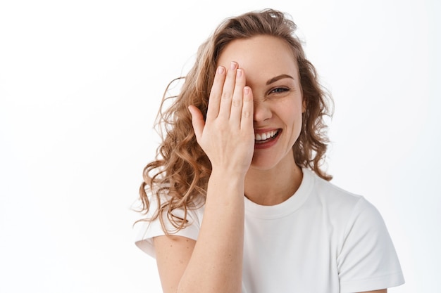 Mulher atraente e cândida esconde metade do rosto atrás da mão e sorri, rindo e mostrando uma expressão natural de rosto feliz, em pé sobre uma parede branca