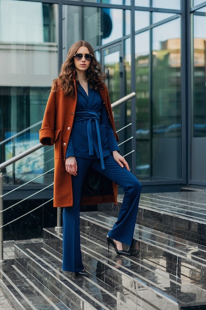 Mulher atraente e elegante com uma caminhada na rua comercial da cidade urbana, vestida com um casaco marrom quente e um terno azul, estilo de rua na moda primavera outono, usando óculos escuros