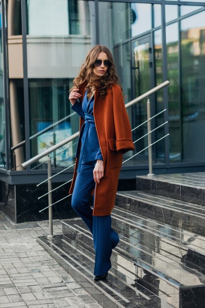 Mulher atraente e elegante com uma caminhada na rua comercial da cidade urbana, vestida com um casaco marrom quente e um terno azul, estilo de rua na moda primavera outono, usando óculos escuros