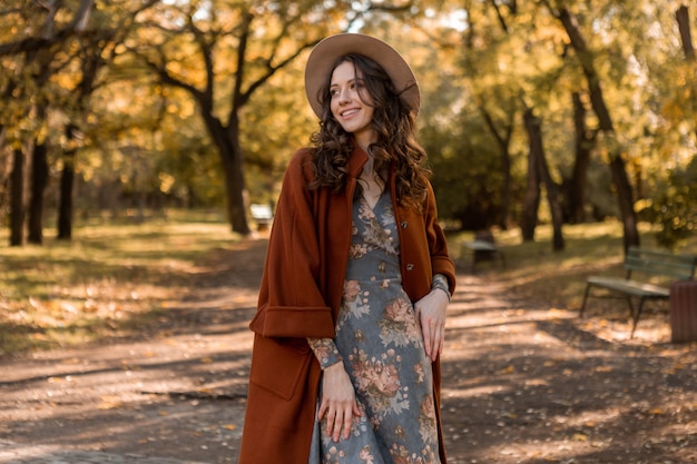 Mulher atraente e elegante e sorridente com cabelos cacheados caminhando no parque vestida com vestido estampado e casaco quente outono na moda, estilo de rua