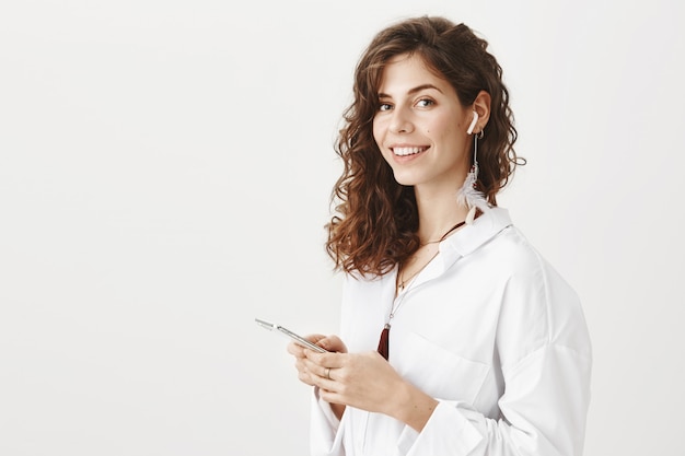 Mulher atraente e elegante ouvindo música em fones de ouvido sem fio, segurando um smartphone