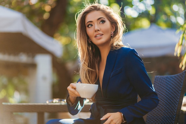Mulher atraente e elegante vestida com um elegante terno azul, sentada à mesa no café, bebendo uma xícara de cappuccino