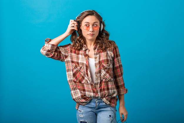 Mulher atraente e engraçada com expressão de surpresa no rosto suspeito olhando de lado ouvindo música em fones de ouvido em camisa quadriculada e jeans isolado no fundo azul do estúdio