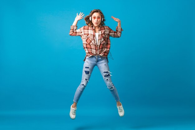 Mulher atraente e engraçada pulando com uma expressão de cara maluca ouvindo música em fones de ouvido em uma camisa quadriculada e calça jeans isolada no fundo azul do estúdio