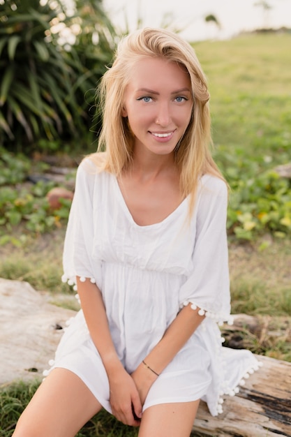 Mulher atraente e esportiva com longos cabelos loiros, grandes olhos azuis e pele limpa, vestida com um vestido branco sentada na madeira